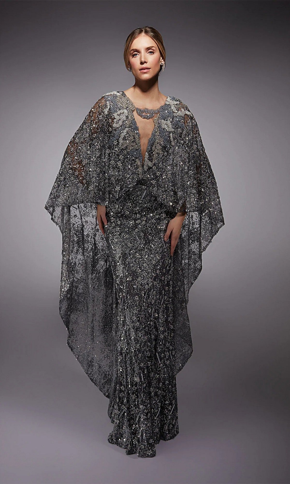 A woman wears the Alyce 27752, a floor-length silver-sequined formal dress with sheer cape-like sleeves, standing against a plain gray background.